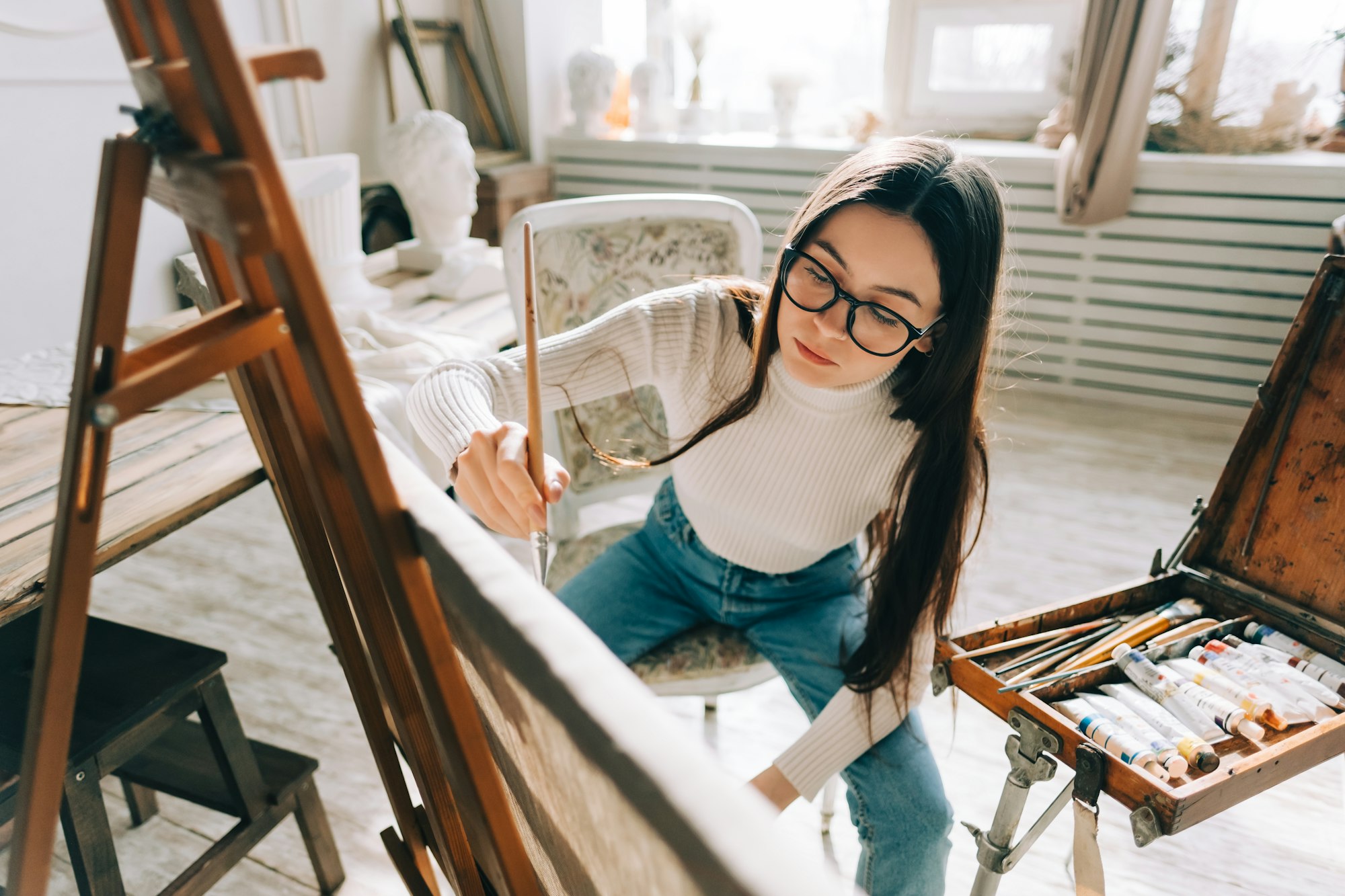 Young caucasian woman artist, holding paintbrush and draws on canvas , working on a home studio.